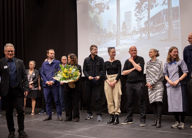 Team Cobe står på scenen i Brønden, en af repræsentanterne for teamet holder en buket med gule blomster. I forggrunden står Borgmester Kent Magelund.
